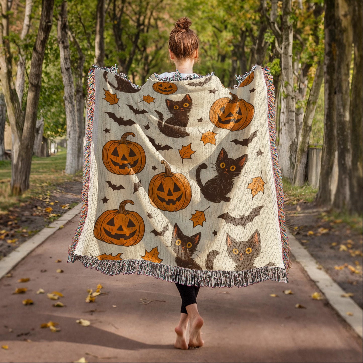 Person walking barefoot down a tree-lined path wearing a Halloween woven blanket featuring black cats, pumpkins, bats, and autumn leaves.